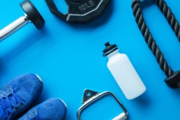 A water bottle, tennis shoes, dumbbell, and jump rope lay on a blue background in a post on how to prevent injury at the gym.