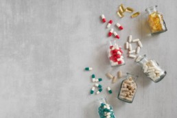 Clear bottles of daily multivitamins are tipped over, spilling colorful capsules onto a concrete surface.