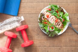 A plate of healthy food, weights, a yoga mat, and a bottle of water on a wooden background, representing different healthy habits.
