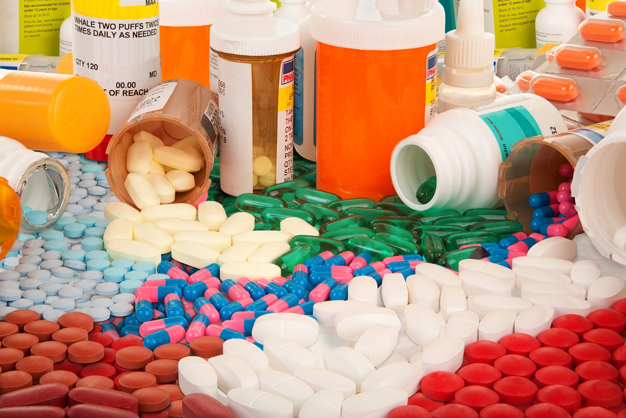 Various sized and shaped pill bottles with assorted medications are spilled over and spread across a white surface.