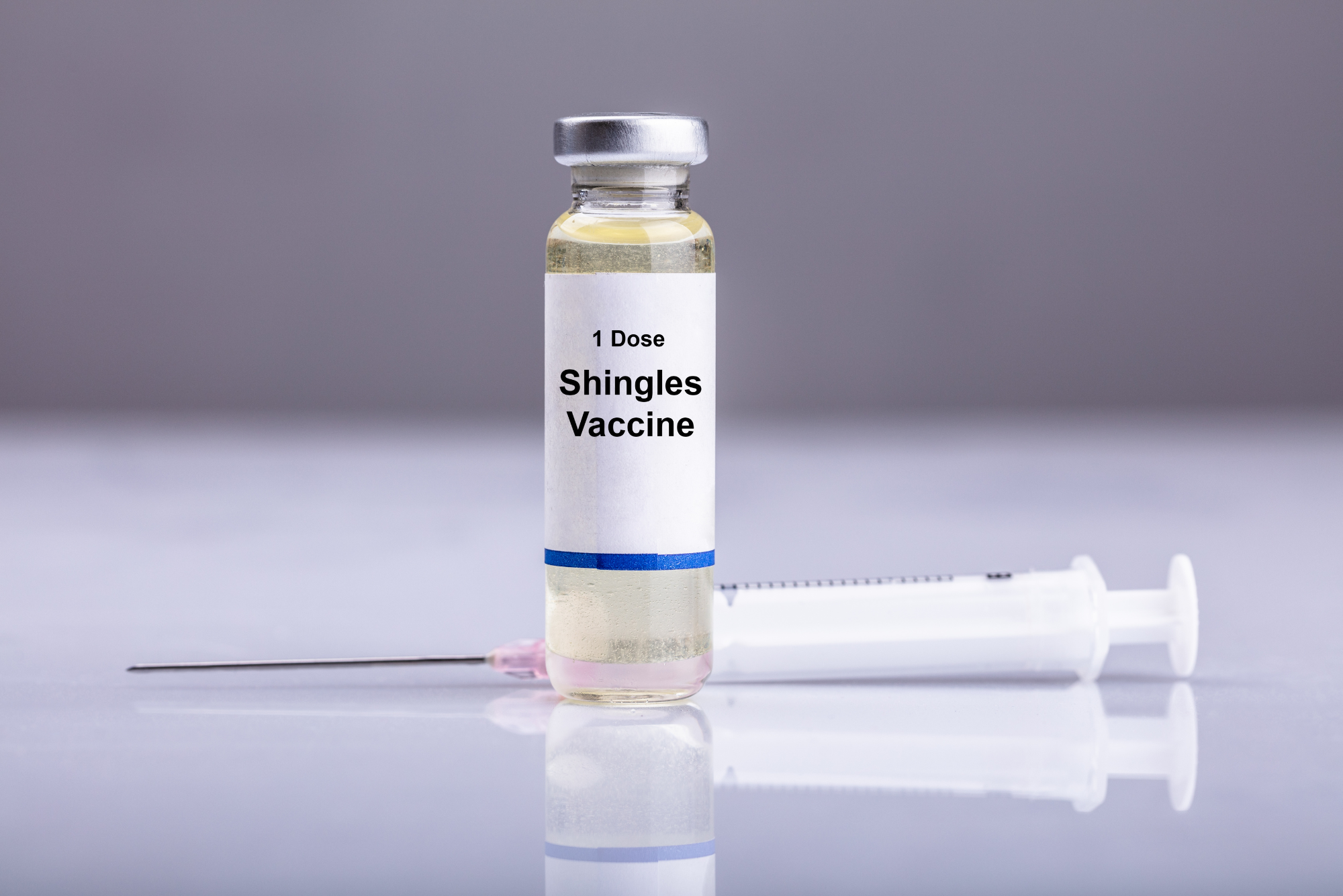 A single dose of the shingles vaccine in a glass vile sits on a clean, white countertop in front of a syringe with a needle attached.