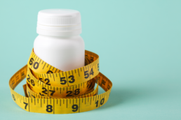 A yellow measuring tape wraps around a white pill bottle, representing evaluating weight loss medication pros and cons.