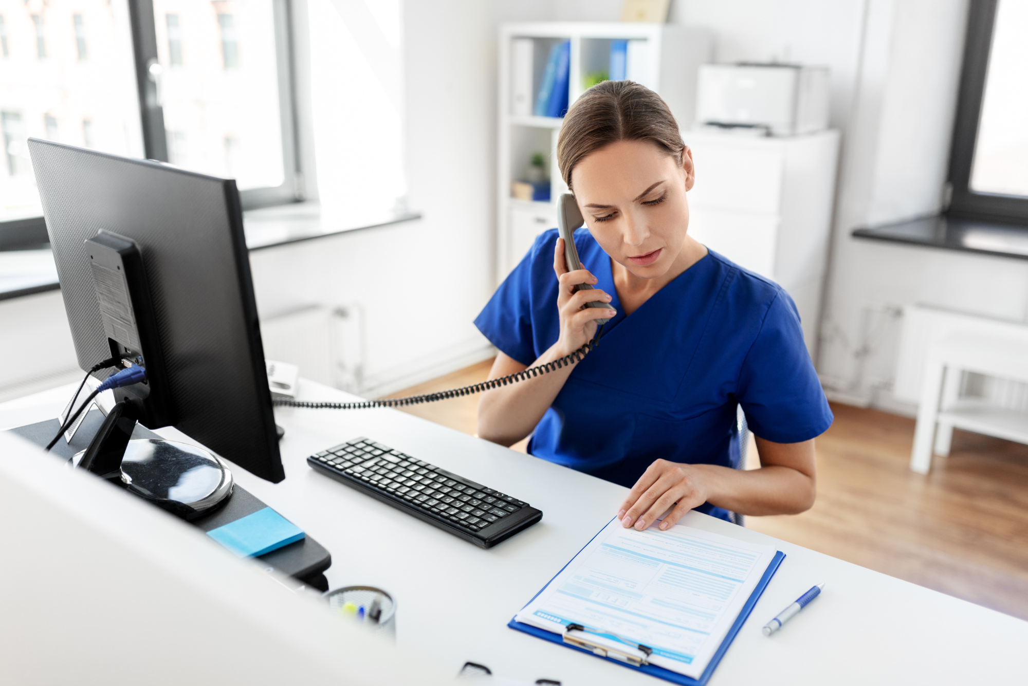 A patient liaison wearing blue scrubs talks on the phone at a white desk.