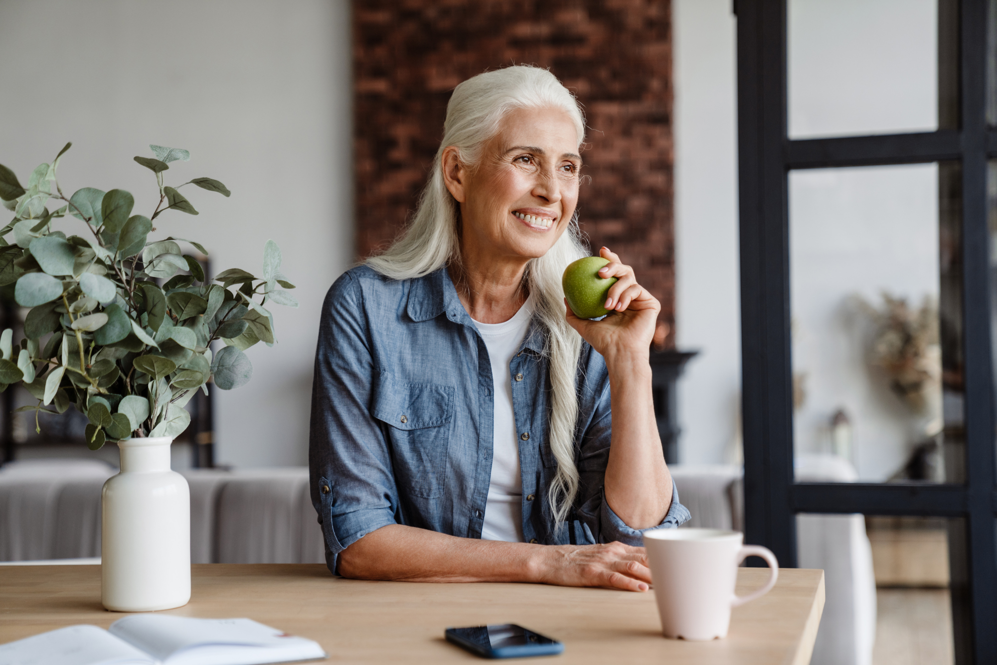 An older woman smiles while eating a green apple because she’s had a long, healthy lifespan.