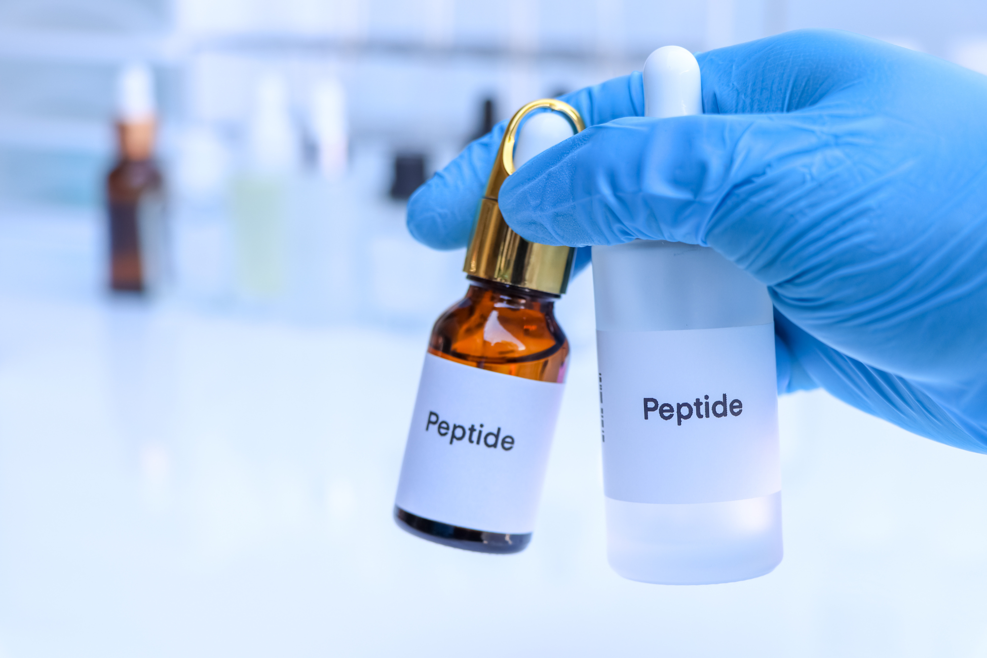 A gloved hand holding two small bottles labeled “Peptide” in a laboratory setting, with a blurred scientific background.