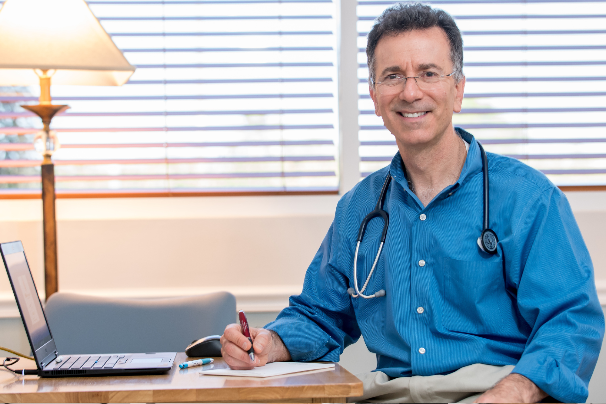Dr. Rosenberg, in a blue shirt with a stethoscope, smiles at his desk with a laptop, pen, and paper in a bright office.