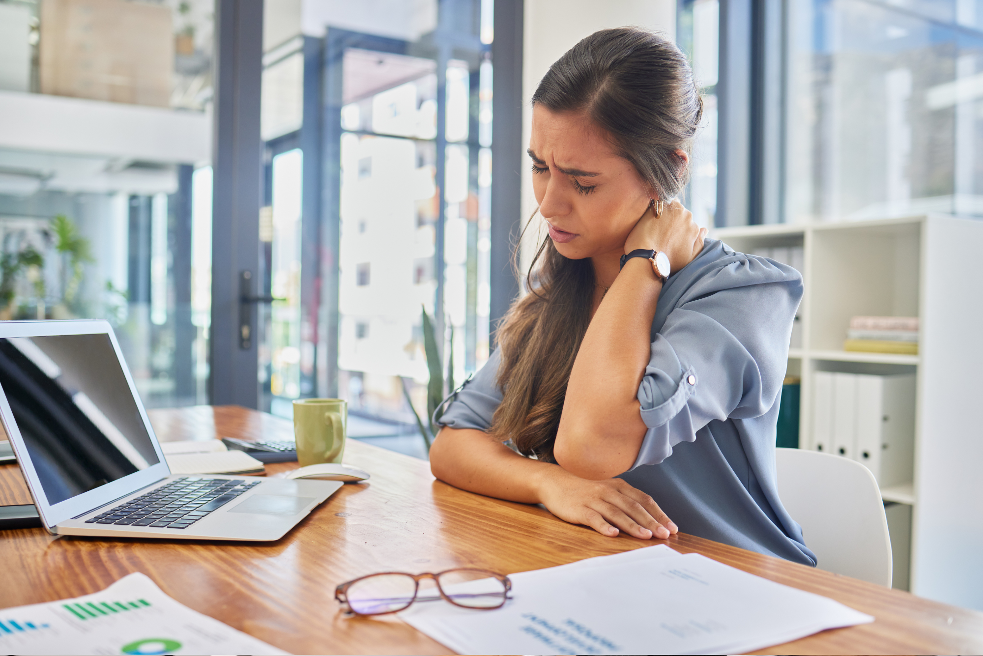 Woman at desk with laptop and papers, holding her neck in pain.