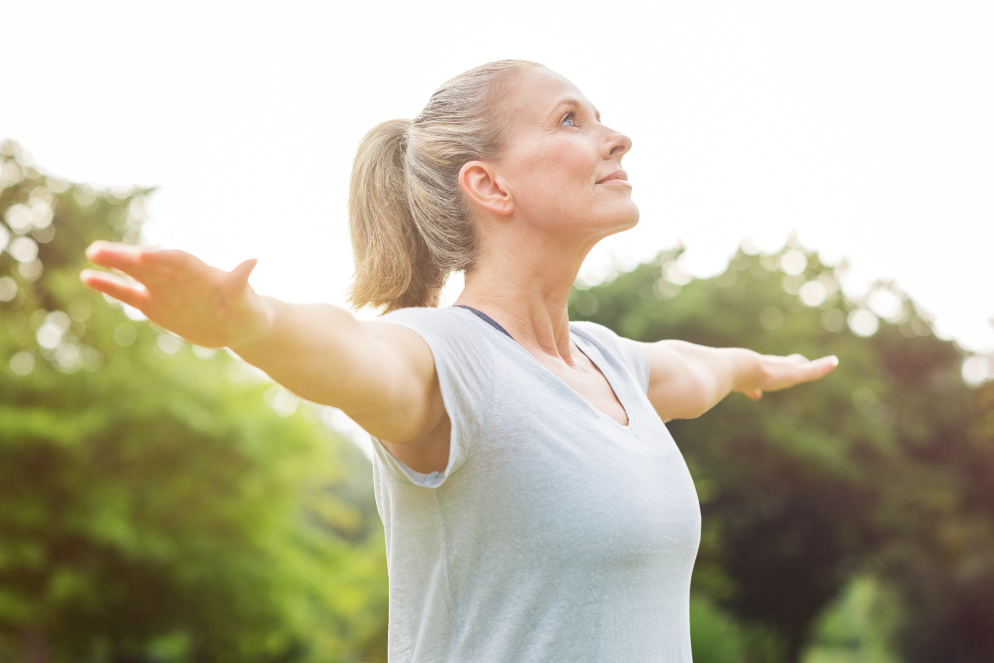 A woman with a ponytail and eyes closed, standing outdoors with arms outstretched.