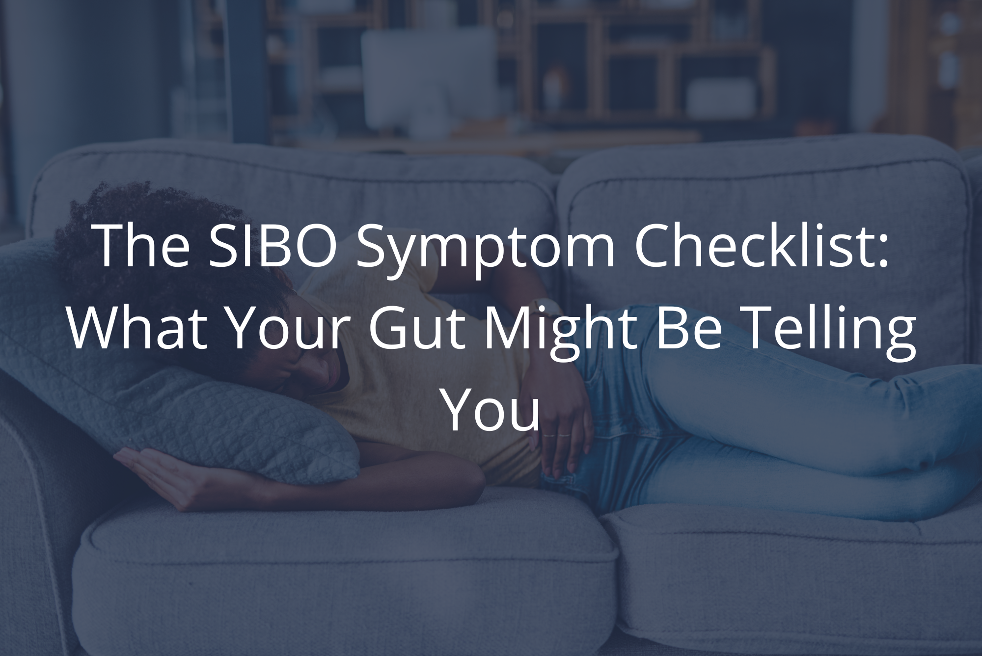 Woman lying on gray couch holding stomach in discomfort, illustrating discomfort often associated with SIBO symptoms.