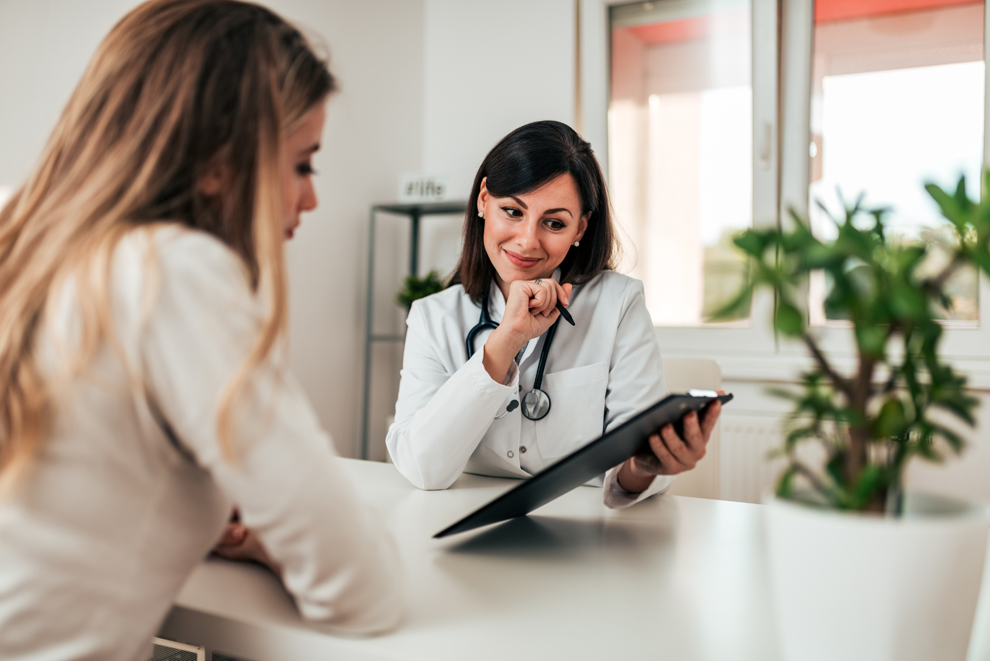 A smiling doctor in a white coat and stethoscope sits across a desk, giving attentive one-on-one time to a patient, illustrating concierge medicine.